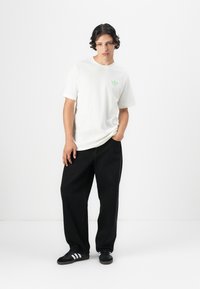 White cotton t-shirt with a green logo, paired with loose black pants and black sneakers featuring white stripes. Simple design and relaxed fit.