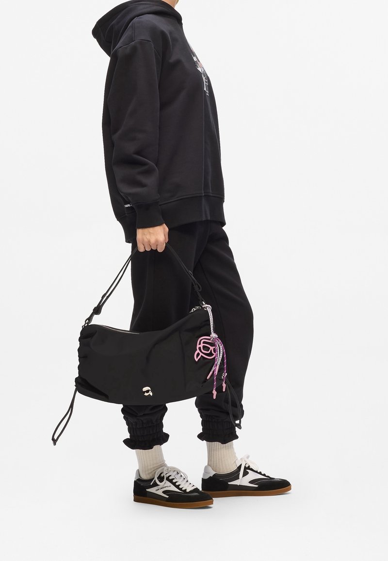 Personne portant un sweat à capuche noir et un pantalon de jogging, tenant un sac à main noir avec logo rose et porte-clés, chaussettes blanches et baskets noires et blanches.