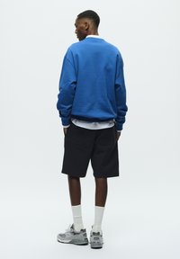 Un sweat-shirt bleu avec des poignets côtelés, associé à un short noir. Des baskets grises et des chaussettes blanches complètent la tenue. Fond blanc uni.