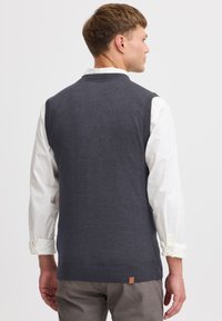 Grauer, gestrickter ärmelloser Pullover mit gerippten Kanten, getragen über einem weißen Langarmshirt; verfügt über ein kleines Logopatch am Saum.