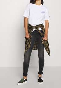 T-shirt blanc avec logo, jean skinny noir, chemise à carreaux verte et noire nouée à la taille, chaussures slip-on camouflage, tissus texturés.