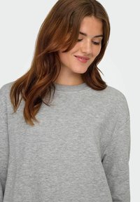 Sudadera gris con un acabado texturizado, cuello redondo y costuras en los hombros caídas. El tejido parece suave y tiene un estilo casual.