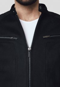 Czarna kurtka zapinana na zamek błyskawiczny z luźnym kołnierzem, dwiema kieszeniami na piersiach z zamkami i gładką teksturą. Design jest minimalistyczny i nowoczesny.