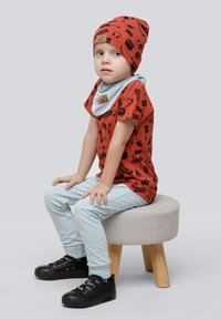 Ein Kind sitzt auf einem Hocker, trägt ein rostfarbenes T-Shirt mit schwarzen grafischen Mustern,hellblaue Hosen und schwarze Turnschuhe.