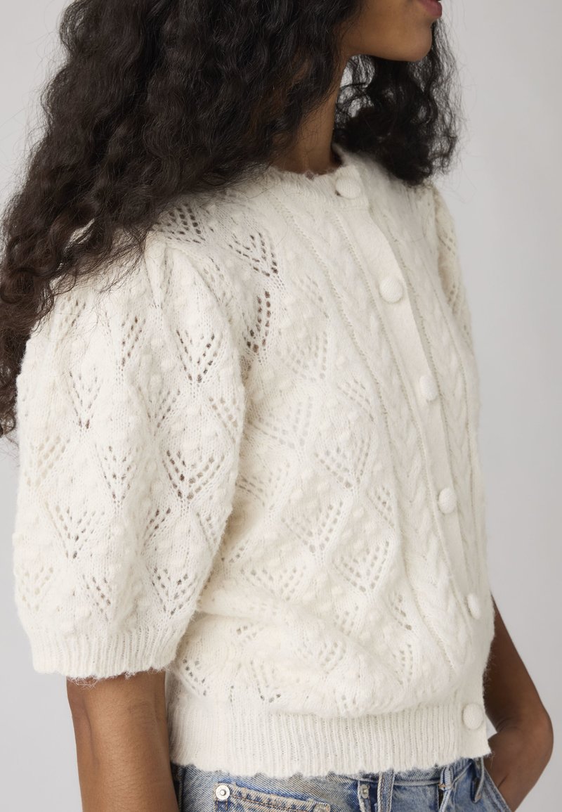 Woman wearing white knit short-sleeve cardigan with button front and textured diamond pattern, paired with light blue denim jeans.