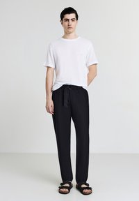 Weiße, strukturierte Kurzarmbluse, lockere schwarze Hose mit gebundenem Gürtel und schwarze Sandalen mit Schleifendetail. Einfaches, neutrales Farbschema.