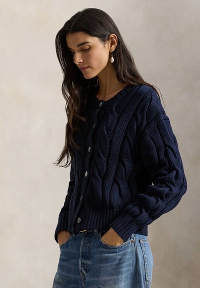 Μπλε σκούρα πλεκτή ζακέτα με σχέδιο κοτσίδα, στρογγυλή λαιμόκοψη και κλείσιμο με κουμπιά. Ριμπ μανσέτες και στρίφωμα αναδεικνύουν την υφή και την εφαρμογή.