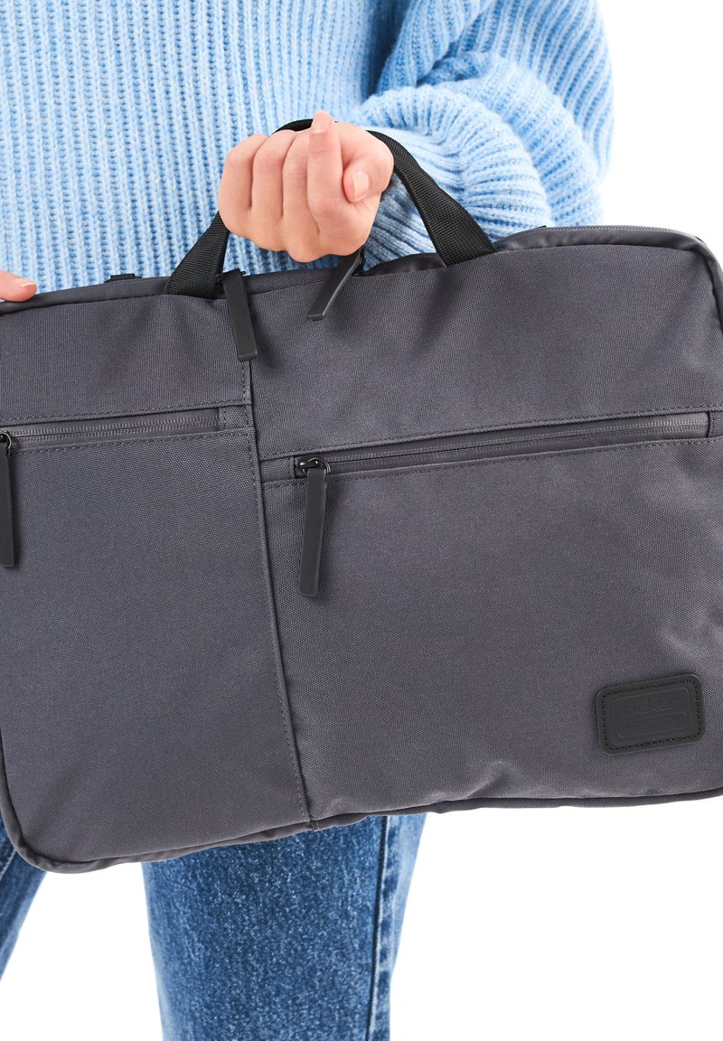 Person in blauer Sweater, der eine graue Stoffakte mit mehreren Reißverschlusstaschen und schwarzen Griffen hält.