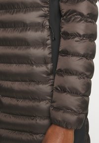 Mörkbrun dunjacka med quiltade sömmar, nylonmaterial och justerbar dragsko vid halslinningen, med en närbild av ärmen och tygtexturen.