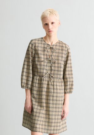 Osoba o krótkich blond włosach, ubrana w beżowo-brązową sukienkę w kratę z trzema wiązaniami z przodu i rękawami do łokcia.