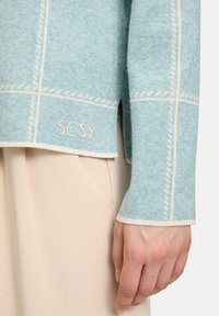 Helles blaues Sweatshirt mit wei�ßer Naht in einem Karomuster, im entspannten Schnitt und einem kleinen "SCSY"-Logo nahe dem Saum.