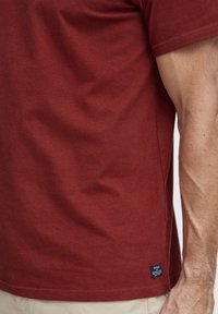Burgundská bavlnená tričko s krátkymi rukávmi. Má kruhový výstrih a malú značkovú etiketu na bočnom šve. Hladká textúra.