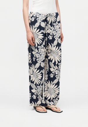 Άτομο που φοράει μαύρο παντελόνι φαρδύ με μεγάλο λευκό ανθικό μοτίβο, σε συνδυασμό με μπεζ σανδάλια τύπου σαγιονάρα και λευκό αμάνικο τοπ.