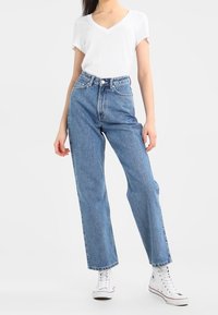 Jeans de mezclilla azules de talle alto con corte recto, combinados con una camiseta blanca de manga corta y escote en pico y zapatillas blancas.