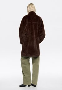 Abrigo de pelo sintético marrón con un cuello alto, fit relajado y mangas largas. Lleva puesto encima de pantalones de pierna ancha verde oliva y zapatos de tacón negros.