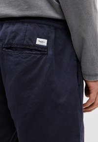 Βαμβακερό παντελόνι σε ναυτικό μπλε χρώμα με πίσω τσέπη που έχει λευκή ετικέτα. Φαίνεται υφασμάτινη υφή και ελαστική μέση. Συμπεριλαμβάνεται γκρι μπλούζα με μακριά μανίκια.