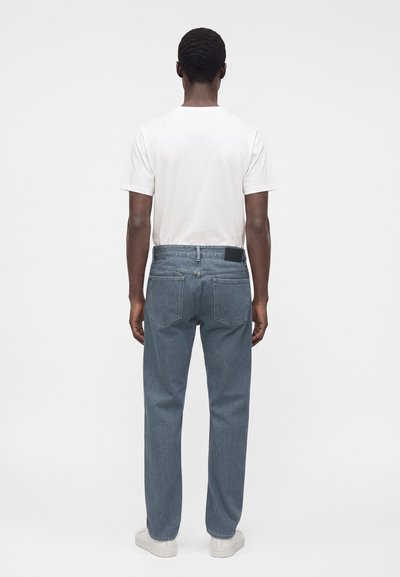 Homme debout de dos, portant une chemise blanche à manches courtes, un jean bleu et des baskets blanches sur un fond blanc uni.