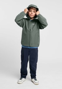 Veste imperméable verte avec capuche, fermeture éclair à l'avant et poignets élastiques. Portée par-dessus une chemise bleue et associée à un pantalon sombre et des baskets.