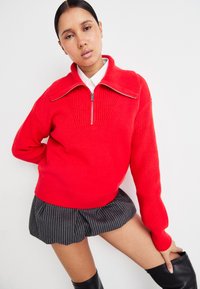 Maglione rosso lavorato a maglia con colletto alto e design a mezza zip, caratterizzato da una texture a coste e dettagli pronunciati sulle spalle, abbinato a una gonna a righe.