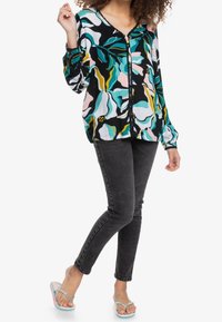 Blusa colorata con un motivo floreale nero, teal, rosa e giallo, abbinata a jeans skinny scuri e infradito multicolori.