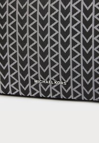 Musträkk mustkott, millel on valge geomeetriline noole muster; sisaldab brändi teksti "MICHAEL KORS" hõbedases metallikvärvis.