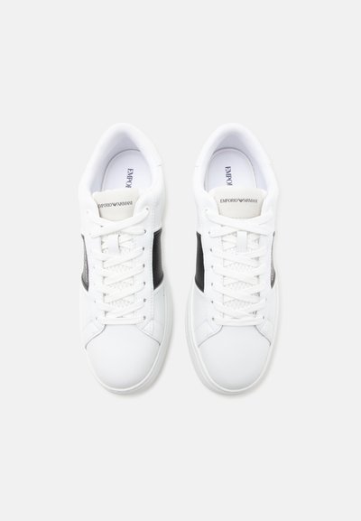 Paire de baskets basses blanches avec des parties latérales noires et des lacets blancs, vues d’en haut sur un fond clair.