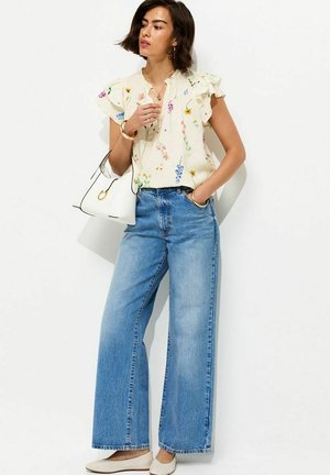 Mujer con blusa crema con flores y mangas con volantes, vaqueros azules de pierna ancha y cintura alta, zapatos planos blancos, sosteniendo bolso blanco, de pie sobre fondo blanco.
