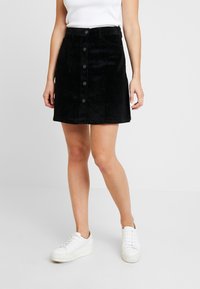 Zwarte corduroy rok met een voorste knoopsluiting, twee zijzakken en een rechte zoom. Gecombineerd met witte sneakers.