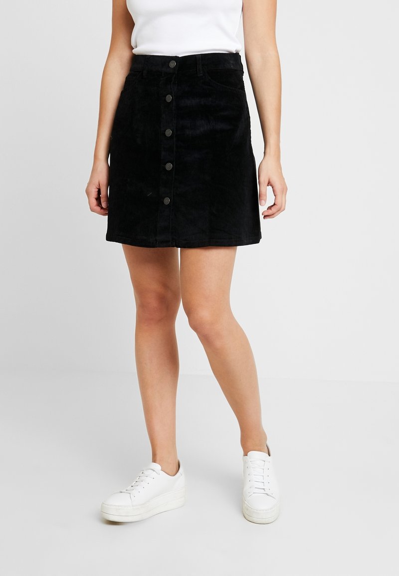Zwarte corduroy rok met een voorste knoopsluiting, twee zijzakken en een rechte zoom. Gecombineerd met witte sneakers.