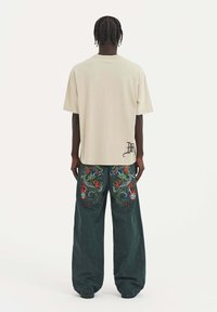 Camiseta oversized beige de mangas cortas, combinada con jeans de pierna ancha verde oscuro que presentan un vibrante bordado de dragón en los bolsillos traseros.