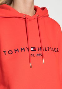 Rode hoodie van zacht materiaal, voorzien van een aansnoerkoord met gouden hardware. Op de voorkant staat het logo "TOMMY HILFIGER" met het jaar 1985 eronder.