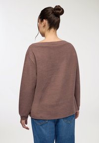 Brązowy, teksturowany sweter z szerokim dekoltem, luźnym krojem i długimi rękawami, noszony z niebieskimi dżinsami. Widok z tyłu.