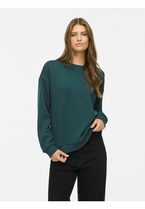 Jeune femme aux longs cheveux bruns portant un pull oversize vert foncé et un pantalon noir, debout sur un fond blanc.
