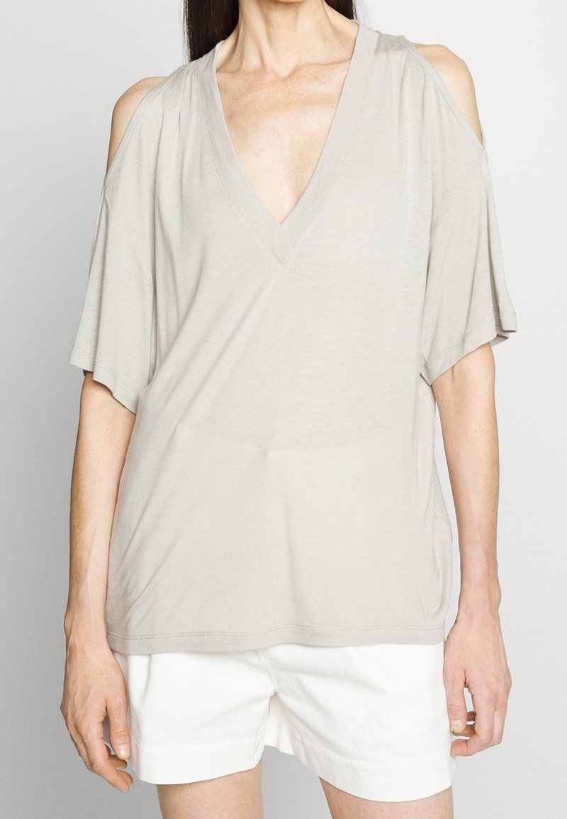 Femme portant un haut beige épaules dénudées en V, associé à un short blanc, debout devant un fond clair uni.
