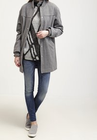Cappotto lungo grigio in misto lana con colletto rotondo e bottoni a scatto, abbinato a un maglione a fantasia, jeans skinny blu e scarpe slip-on grigie.