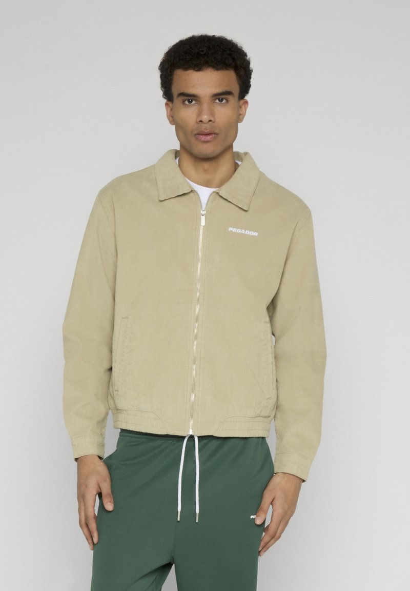 Hombre con chaqueta beige con cremallera y pantalones verdes con cordón, posando contra un fondo claro y liso.