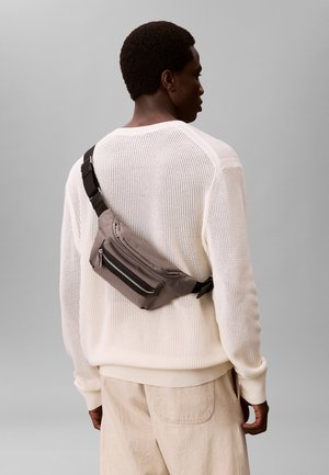 Mand iført beige bukser og hvid strikket sweater med grå crossbody talletaske mod en ensfarvet grå baggrund.