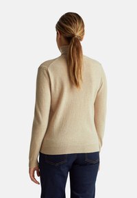 Beżowy sweter z dzianiny z wysokim kołnierzem, ściągaczami przy mankietach i u dołu. Miękka faktura, dopasowany krój i gładkie wykończenie, noszony z ciemnoniebieskimi dżinsami.