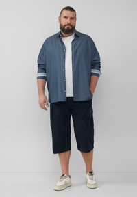 Niebieska koszula z długim rękawem z podwiniętymi rękawami, biała koszulka pod spodem, granatowe szorty cargo i kremowe sneakersy. Casualowy, luźny krój.