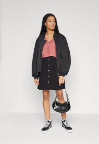 Zwarte bomberjack, roze top, zwarte knoopskirt, zwarte sneakers en een kleine zwarte handtas. Gladde texturen en casual stijl.