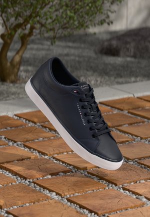 Schwarzer Tommy Hilfiger Sneaker mit weißer Sohle, der über einem Boden mit Holz- und Kieselmuster im Freien schwebt.