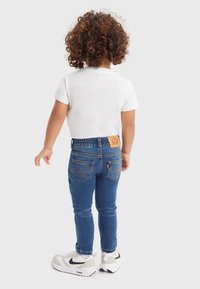 Jeans de mezclilla azul con un ajuste ceñido, cinturilla elástica y bolsillos traseros, combinados con una camiseta blanca de manga corta y zapatillas deportivas.