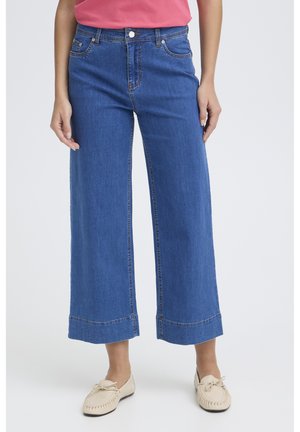 Žena oblečená v modrých širokých zkrácených džínách, růžovém topu a béžových mokasínách, stojící na jednobarevném světlém pozadí.