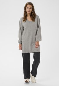 Šedá tunika s dlouhým rukávem a výstřihem do V, disponující měkkou texturou. Spárována s černými džíny rovného střihu a bílými teniskami.