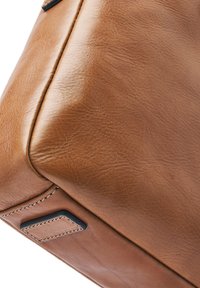 Close-up of the bottom corner of a tan leather bag showing detailed stitching and a small black-edged leather patch.