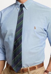 Camicia azzurro chiaro con bottone e un piccolo logo polo. Cravatta a righe blu navy e verde con accenti dorati. Cintura in pelle marrone. Fit su misura.