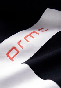 Musta kangas, jossa on näkyvä valkoinen etiketti, jossa on kohokuvioitu punainen teksti "prmc". Pintakuvio on sileä, mikä korostaa värien välistä kontrastia.