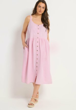 Kobieta w różowej, bezrękawnikowej, midi sukience na guziki z kieszeniami, w beżowych sandałach na koturnie, stojąca na tle jednolitego tła.