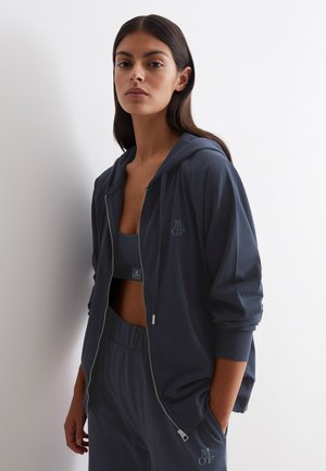 Haut de survêtement zippé bleu marine avec une coupe décontractée, fabriqué en tissu doux. Présente une capuche, des poches latérales et un détail de logo sur la poitrine et la taille.