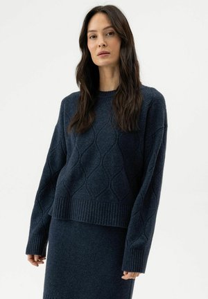 Femme aux longs cheveux foncés portant un pull à torsades bleu foncé et une jupe assortie, debout devant un fond blanc uni.